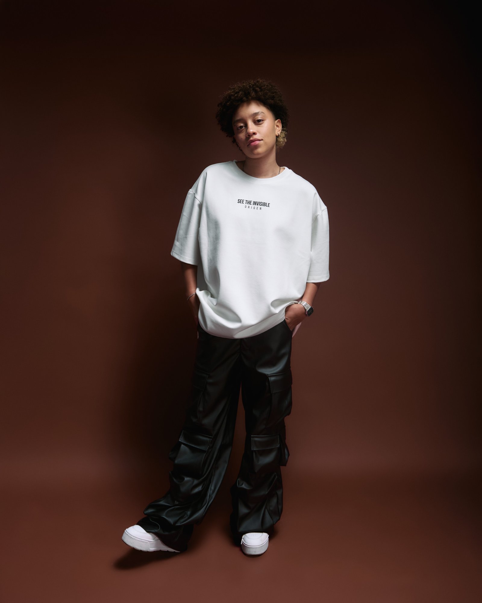 Youthful woman wearing a white oversized T-shirt with the text "See The Invisible" and black cargo pants, standing confidently against a brown gradient background, showcasing casual streetwear fashion.