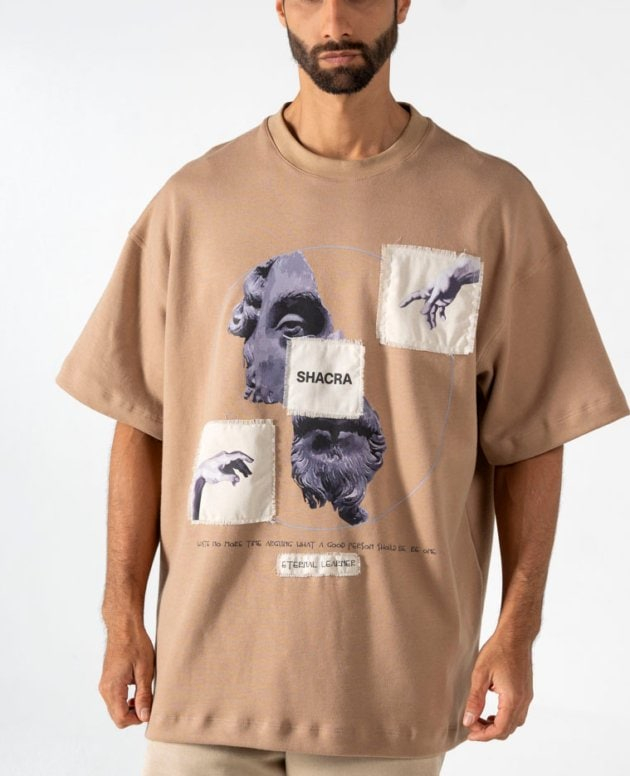A man wearing a beige graphic T-shirt with artistic face and hand illustrations, featuring the brand name Shacra, against a plain gray background.