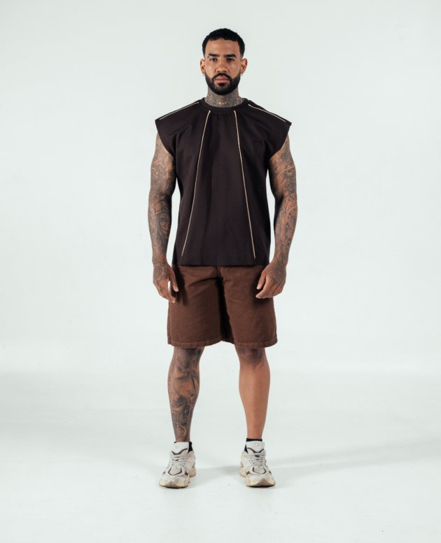 Bold tattooed man wearing a black sleeveless top with gold accents, brown shorts, and athletic sneakers, standing confidently in a minimal studio setting, showcasing modern streetwear fashion.