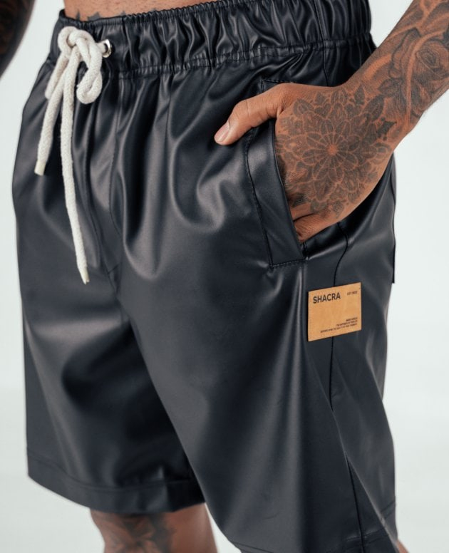 Black faux leather shorts with a drawstring waist and a small leather patch label, worn by a model with tattoos on their hand and leg. Perfect for stylish streetwear and casual summer outfits.