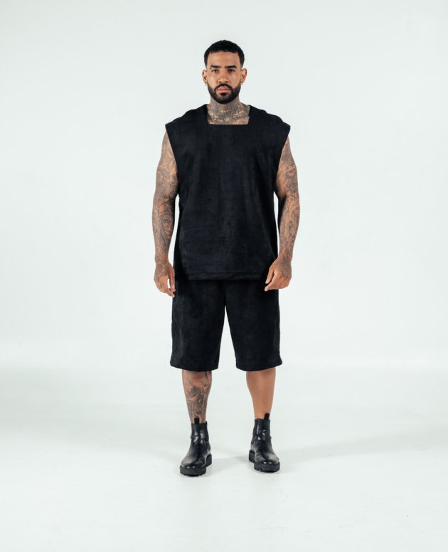 Bold tattooed man wearing black sleeveless top, shorts, and combat boots in studio against white background, showcasing edgy fashion style and body art.